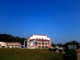 A modern building with an angular, multi-story design sits in the foreground, framed by a vast green lawn and lush trees in the background. The sky is clear and blue, contributing to a peaceful ambiance. Large letters spelling 'IITG' are visible in the landscape.