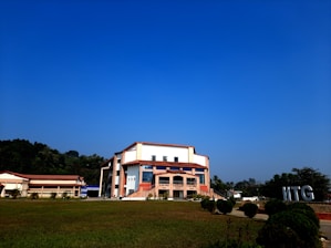 A modern building with an angular, multi-story design sits in the foreground, framed by a vast green lawn and lush trees in the background. The sky is clear and blue, contributing to a peaceful ambiance. Large letters spelling 'IITG' are visible in the landscape.