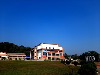 A modern building with an angular, multi-story design sits in the foreground, framed by a vast green lawn and lush trees in the background. The sky is clear and blue, contributing to a peaceful ambiance. Large letters spelling 'IITG' are visible in the landscape.
