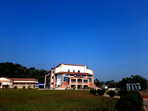 A modern building with an angular, multi-story design sits in the foreground, framed by a vast green lawn and lush trees in the background. The sky is clear and blue, contributing to a peaceful ambiance. Large letters spelling 'IITG' are visible in the landscape.