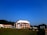 A modern building with an angular, multi-story design sits in the foreground, framed by a vast green lawn and lush trees in the background. The sky is clear and blue, contributing to a peaceful ambiance. Large letters spelling 'IITG' are visible in the landscape.