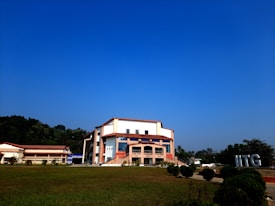 A modern building with an angular, multi-story design sits in the foreground, framed by a vast green lawn and lush trees in the background. The sky is clear and blue, contributing to a peaceful ambiance. Large letters spelling 'IITG' are visible in the landscape.