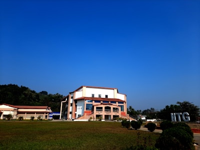 A modern building with an angular, multi-story design sits in the foreground, framed by a vast green lawn and lush trees in the background. The sky is clear and blue, contributing to a peaceful ambiance. Large letters spelling 'IITG' are visible in the landscape.