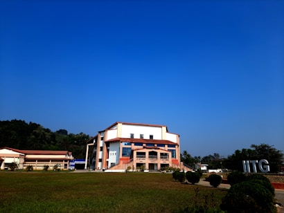 A modern building with an angular, multi-story design sits in the foreground, framed by a vast green lawn and lush trees in the background. The sky is clear and blue, contributing to a peaceful ambiance. Large letters spelling 'IITG' are visible in the landscape.