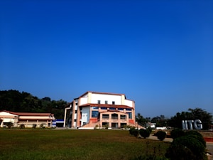 A modern building with an angular, multi-story design sits in the foreground, framed by a vast green lawn and lush trees in the background. The sky is clear and blue, contributing to a peaceful ambiance. Large letters spelling 'IITG' are visible in the landscape.