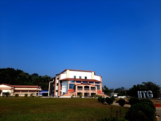 A modern building with an angular, multi-story design sits in the foreground, framed by a vast green lawn and lush trees in the background. The sky is clear and blue, contributing to a peaceful ambiance. Large letters spelling 'IITG' are visible in the landscape.