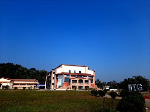 A modern building with an angular, multi-story design sits in the foreground, framed by a vast green lawn and lush trees in the background. The sky is clear and blue, contributing to a peaceful ambiance. Large letters spelling 'IITG' are visible in the landscape.