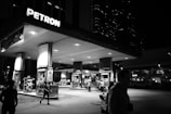 A gas station illuminated at night, with several people visible in the foreground. The station is branded Petron and features multiple fuel pumps and signs. There are structures in the background, including tall buildings with lights on, suggesting an urban setting.