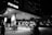 A gas station illuminated at night, with several people visible in the foreground. The station is branded Petron and features multiple fuel pumps and signs. There are structures in the background, including tall buildings with lights on, suggesting an urban setting.