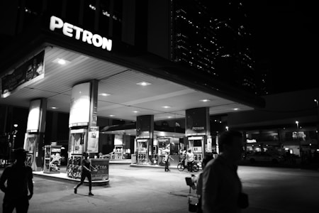 A gas station illuminated at night, with several people visible in the foreground. The station is branded Petron and features multiple fuel pumps and signs. There are structures in the background, including tall buildings with lights on, suggesting an urban setting.