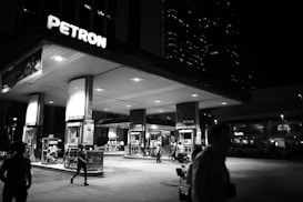 A gas station illuminated at night, with several people visible in the foreground. The station is branded Petron and features multiple fuel pumps and signs. There are structures in the background, including tall buildings with lights on, suggesting an urban setting.