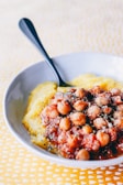 A white bowl containing a dish of chickpeas cooked in a red tomato-based sauce, garnished with herbs, served over a bed of creamy yellow polenta. A black spoon is placed on the side of the bowl. The background consists of a patterned yellow tablecloth.