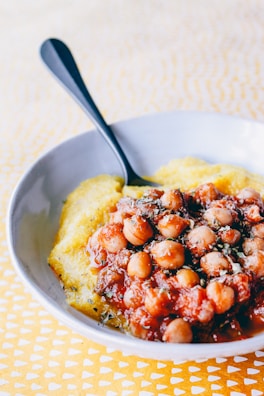 A white bowl containing a dish of chickpeas cooked in a red tomato-based sauce, garnished with herbs, served over a bed of creamy yellow polenta. A black spoon is placed on the side of the bowl. The background consists of a patterned yellow tablecloth.