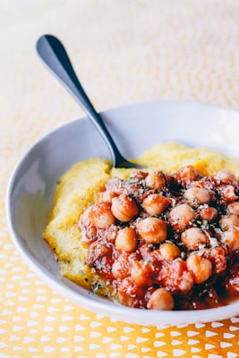 A white bowl containing a dish of chickpeas cooked in a red tomato-based sauce, garnished with herbs, served over a bed of creamy yellow polenta. A black spoon is placed on the side of the bowl. The background consists of a patterned yellow tablecloth.
