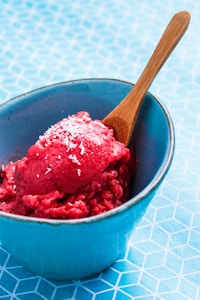 A bright red, textured frozen dessert topped with shredded coconut sits in a light blue bowl. A wooden spoon is placed into the dessert, and the bowl is on a geometric patterned surface.