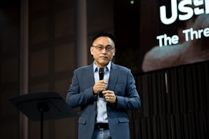 A person wearing glasses, dressed in a blue suit and white shirt, is holding a microphone and standing on a stage. Behind them, a large screen displays partially visible text. The setting appears to be a formal event or presentation.