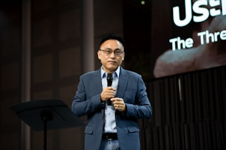 A person wearing glasses, dressed in a blue suit and white shirt, is holding a microphone and standing on a stage. Behind them, a large screen displays partially visible text. The setting appears to be a formal event or presentation.