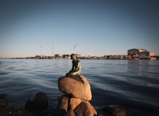 A small bronze statue of a mermaid sits on a rock by the water's edge, gazing towards the horizon. The scene is set against a calm water backdrop with industrial buildings and structures in the distance. Wind turbines can be seen on the horizon under a clear blue sky.