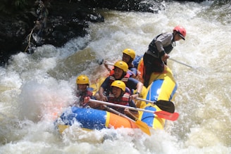 A group of people wearing helmets and life jackets are paddling a colorful inflatable raft through turbulent white-water rapids. Water splashes around them as they navigate the river with focused expressions.