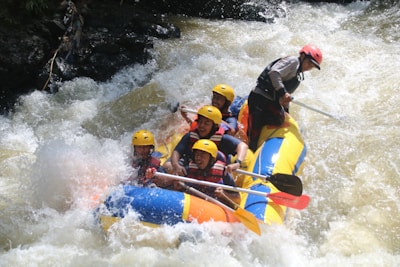 A group of people wearing helmets and life jackets are paddling a colorful inflatable raft through turbulent white-water rapids. Water splashes around them as they navigate the river with focused expressions.