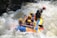 A group of people wearing helmets and life jackets are paddling a colorful inflatable raft through turbulent white-water rapids. Water splashes around them as they navigate the river with focused expressions.