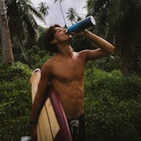 A shirtless person with a fit physique is holding a surfboard under one arm while drinking from a blue bottle. They are standing in a lush, tropical environment with tall palm trees in the background.
