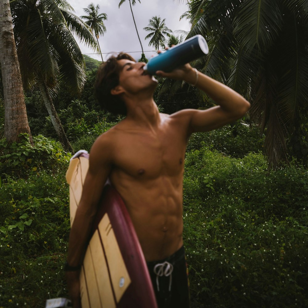 a shirtless man holding a surfboard and drinking from a bottle