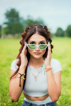 A young woman with long brown hair stands in a lush green field. She is wearing a white crop top and blue jeans, accessorized with colorful bracelets and a necklace. Her hair is styled with small colorful hair ties. She is holding a pair of light green sunglasses, emphasizing a playful and stylish look.