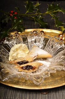 A decorative glass plate holds crispy pastry filled with dark fruit filling, dusted with powdered sugar, accompanied by a dollop of cream. The setup is enhanced by a golden tray underneath and sprigs of holly with bright red berries as background decor.