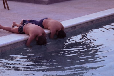Two children are lying on their stomachs at the edge of a swimming pool with their heads leaning over the side, appearing to look into the water. The setting appears to be outdoors on a concrete or tiled pool deck.