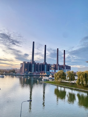 A large industrial building with four tall chimneys is situated beside a reflective body of water. The scene is framed by greenery, including trees and grassy areas. The sky overhead is expansive and partly cloudy, illuminated with soft, natural light, suggesting either early morning or late afternoon.
