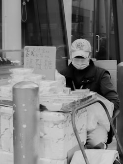 A person wearing a cap and face mask sits behind a street vendor stall, surrounded by boxes and packaged goods with a sign displaying text in Chinese. They are likely involved in selling or managing the items.