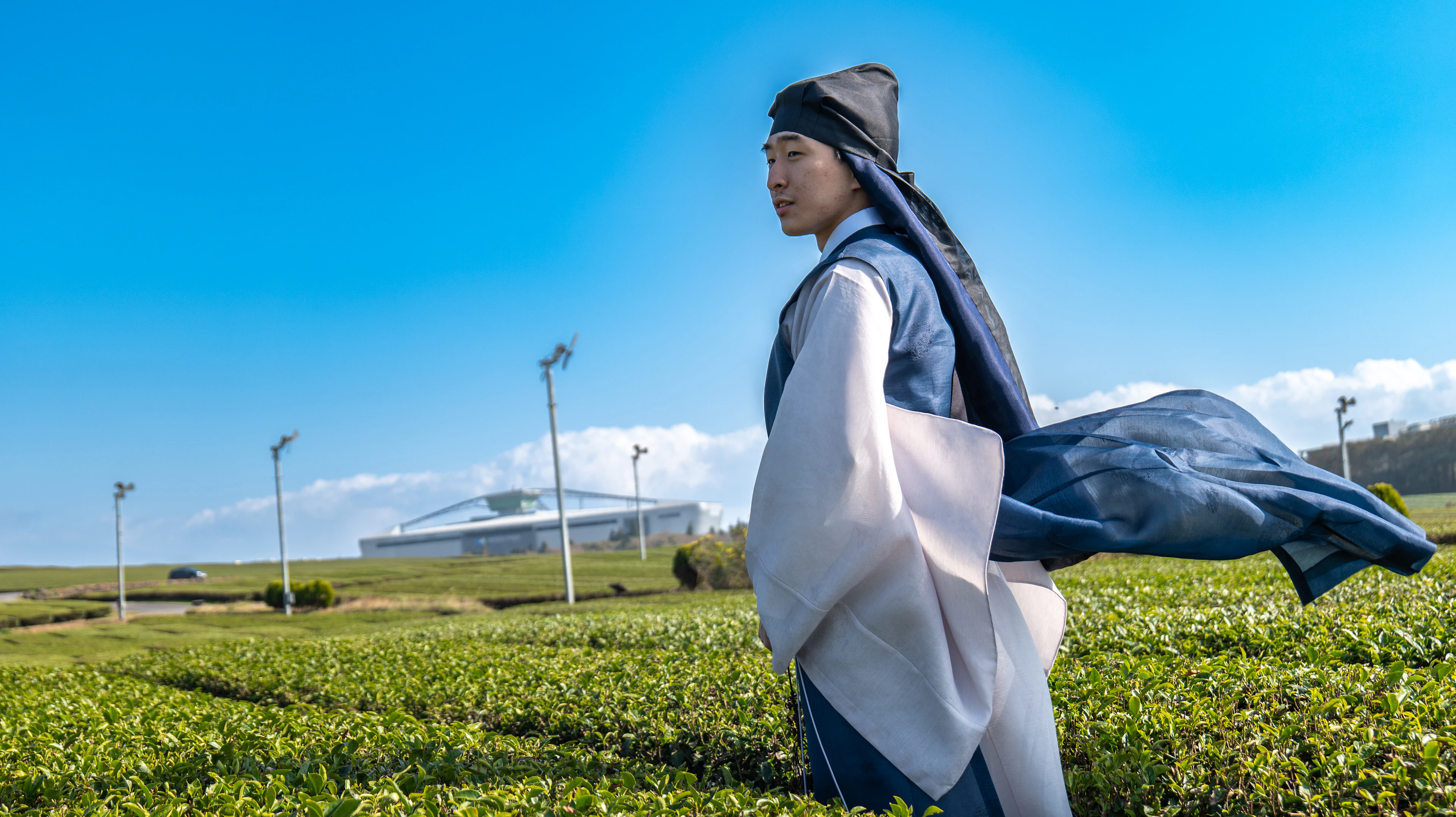 A woman in a flowing hanbok-style dress walks through a green tea field with wind turbines behind her.