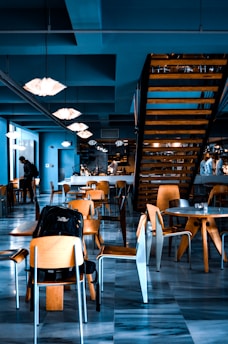 A modern cafeteria interior with wooden tables and chairs arranged in an open seating layout. The ceiling has geometric pendant lights, and a prominent wooden staircase is visible. Some people are present in the background, contributing to a lively atmosphere.