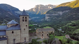 A historic stone building with a bell tower stands in the foreground, surrounded by lush greenery and set against a backdrop of majestic mountains bathed in sunlight. The landscape features a valley with scattered trees and small houses, conveying a serene and peaceful rural setting.