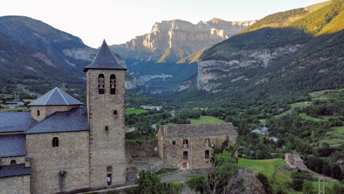 A historic stone building with a bell tower stands in the foreground, surrounded by lush greenery and set against a backdrop of majestic mountains bathed in sunlight. The landscape features a valley with scattered trees and small houses, conveying a serene and peaceful rural setting.
