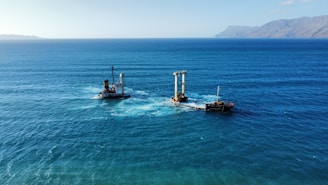 A large body of water with a ship partially submerged and a visible structure with tall pillars. The ocean is expansive, with a distant view of a mountainous coastline under a clear blue sky.