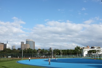 A bright outdoor sports complex features a blue running track, surrounded by green grass and trees. Several people are visible, engaged in activities on the track. In the background, urban high-rise buildings rise against a partly cloudy sky.