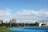 A bright outdoor sports complex features a blue running track, surrounded by green grass and trees. Several people are visible, engaged in activities on the track. In the background, urban high-rise buildings rise against a partly cloudy sky.
