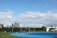 A bright outdoor sports complex features a blue running track, surrounded by green grass and trees. Several people are visible, engaged in activities on the track. In the background, urban high-rise buildings rise against a partly cloudy sky.