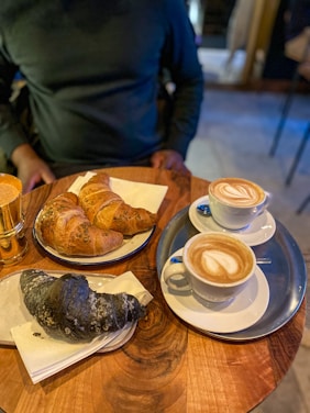 A wooden table holds a variety of pastries and two cups of coffee. Two croissants on a blue-rimmed plate and a unique black croissant on a separate plate are arranged neatly. The coffee cups feature latte art on their creamy surfaces, placed on saucers with a silver tray underneath.
