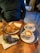 A wooden table holds a variety of pastries and two cups of coffee. Two croissants on a blue-rimmed plate and a unique black croissant on a separate plate are arranged neatly. The coffee cups feature latte art on their creamy surfaces, placed on saucers with a silver tray underneath.