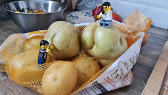 A bag of potatoes sits on a kitchen counter. Two small figures resembling toy construction workers are playfully positioned on the potatoes. A metal bowl with tomatoes and some kitchen utensils are visible in the background.