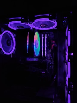 A computer casing interior is illuminated with vibrant RGB lighting. Multiple fans are visible with purple and blue LED accents. The motherboard and RAM sticks are discernible, along with various cables and hardware components.