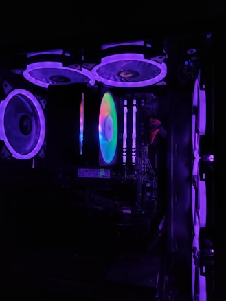 A computer casing interior is illuminated with vibrant RGB lighting. Multiple fans are visible with purple and blue LED accents. The motherboard and RAM sticks are discernible, along with various cables and hardware components.