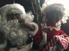 Two festive figures featuring a Santa and Mrs. Claus are dressed in traditional holiday attire. Santa has a curly white beard and wears a red hat with a white trim, while Mrs. Claus dons a bonnet and is holding a striped stocking and a candle. The scene appears to be in a dimly lit room.
