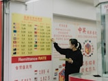 A person wearing a mask stands in front of a currency exchange board displaying various exchange rates between different currencies such as USD, EUR, JPY, and more. The board is yellow with red and black text, accompanied by several national flags. The person appears to be holding a small tray and pointing at the board, likely indicating or discussing the rates. The setting seems to be inside a financial service office, with additional informational posters in the background written in Chinese and English.
