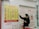 A person wearing a mask stands in front of a currency exchange board displaying various exchange rates between different currencies such as USD, EUR, JPY, and more. The board is yellow with red and black text, accompanied by several national flags. The person appears to be holding a small tray and pointing at the board, likely indicating or discussing the rates. The setting seems to be inside a financial service office, with additional informational posters in the background written in Chinese and English.