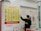 A person wearing a mask stands in front of a currency exchange board displaying various exchange rates between different currencies such as USD, EUR, JPY, and more. The board is yellow with red and black text, accompanied by several national flags. The person appears to be holding a small tray and pointing at the board, likely indicating or discussing the rates. The setting seems to be inside a financial service office, with additional informational posters in the background written in Chinese and English.