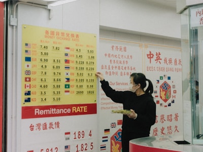A person wearing a mask stands in front of a currency exchange board displaying various exchange rates between different currencies such as USD, EUR, JPY, and more. The board is yellow with red and black text, accompanied by several national flags. The person appears to be holding a small tray and pointing at the board, likely indicating or discussing the rates. The setting seems to be inside a financial service office, with additional informational posters in the background written in Chinese and English.